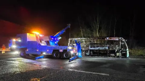 The burned out shell of a coach on the side of a motorway slip road. It is attached to a grey towing vehicle.