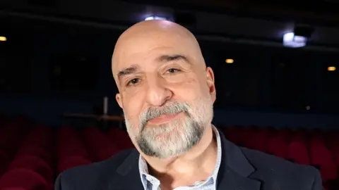 Qays Najm/BBC Comedian and actor Omid Djalili smiles at the camera while standing in the Ipswich Regent Theatre. He is bald and has a grey goatee beard. He is wearing a navy suit jacket with a blue shirt. Red seats of the theatre can be seen behind him.