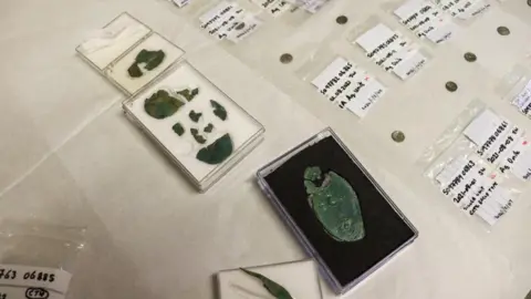 Cotswold District Council Fragments of miniature iron and copper-alloy shields and spearheads on a table in cases next to rows of coins with labels