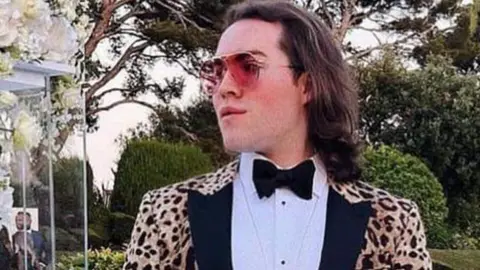 Jack Watkin is holding a handbag while wearing a leopard print jacket, white shirt and black bow tie. He is also wearing sunglasses in this social media image taken in a landscaped garden with large pond.