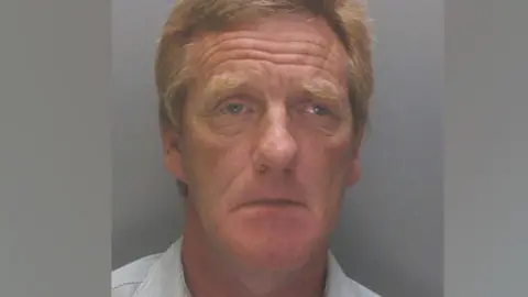 A police mugshot of Anthony Burns, 63, who has ginger hair and is wearing a light blue collared shirt. He is looking into the camera with a sorrowful expression. 