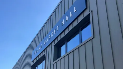 the outside of a building. It's modern looking, with overlapping grey vertical slates. Above two small windows a black sign reads "COLNE MARKET HALL"