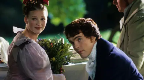 Joanna Page and Ciaran McMenamin as David Copperfield. Page is wearing a light pink dress while McMenamin has a blue velvet jacket with a white shirt underneath. He has short brown curly hair.