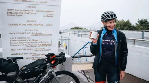 Cyclist Christina MacKenzie to the right of screen in black cycling attire, holding a her winning silver hip flask filled with irn-bru. Her grey Liv bike is to the left of screen. Photo taken at South Queensferry Memorial. 