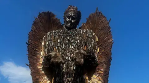The Knife Angel statue stands against a blue sky. It looks like a man with his hands outstretched, palms up. He has wings and is made out of knives.