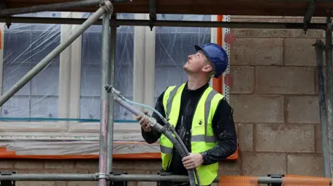 Reuters A construction worker wearing a blue helmet and a yellow high-viz jacket works on scaffolding outside a building.