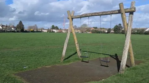 A wood-framed child's swing in a green and unmarked playing field. The frame supports two black plastic swing seats on metal chains. The large field is otherwise featureless. Beyond, there are houses on the horizon and a large orange crane.