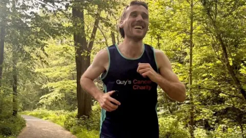 Ollie Nash A man smiling whilst running along a wooded path in image (87).jpg, wearing a dark blue vest featuring the text "Guy's Cancer Charity".