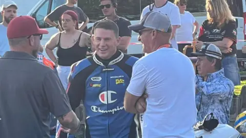 Armina Magaway-Alih Channing Tatum is wearing blue and black leathers and is laughing. There are two gentleman facing him either side, and a crowd of people behind him. 