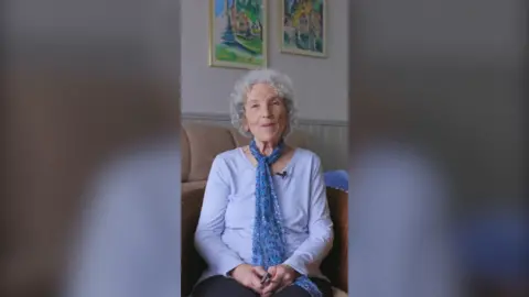 An older woman with curly grey hair, sits in an armchair looking at the camera. She's wearing a lilac top and a blue scarf.