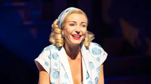 Trafalgar Entertainment Helen George wears a blue and white top and head scarf, with curly blonde hair and lipstick. She's smiling