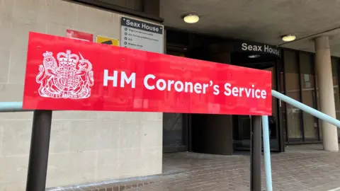 Lewis Adams/BBC A red sign that says HM Coroner's Service. It is at the entrance to Essex Coroner's Court, which is a cream building that has a black revolving door and brick paving outside.