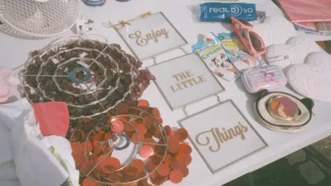 Paul Blakemore A car boot sale stand with three square signs in a row reading 'Enjoy the little things' with other random objects scattered across the table. 