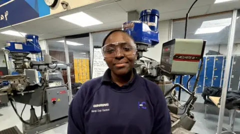 A girl wearing safety glasses with her hair tied back out of shot also wearing a navy blue sweatshirt. She is stood in a factory environment with two large machines behind her.