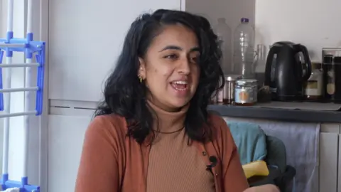 A woman with black hair and olive skin sits at a table and behind her a kettle is visible on the counter