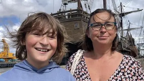 Robby West/BBC Cameron and his Mum, April. They are standing in front of the ship docked at Great Yarmouth. Cameron is wearing a light blue hoodie and is looking directly at the camera and smiling. April is wearing a black floral dress and glasses, and is looking directly at the camera while smiling.