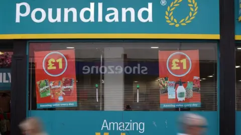 A Poundland store shop front, with two people, blurred, walking past. There are signs in the window that say £1. 