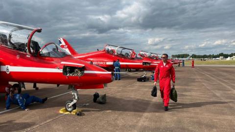 The Red Arrows on the tarmac at RAF Fairford, with a pilot walking in front of them holding two backpacks