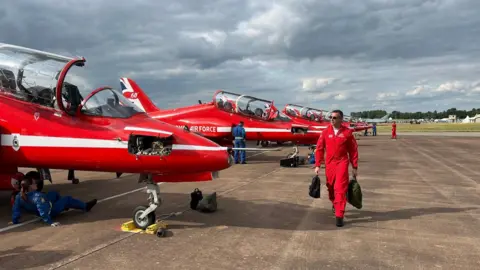 The Red Arrows on the tarmac at RAF Fairford, with a pilot walking in front of them holding two backpacks