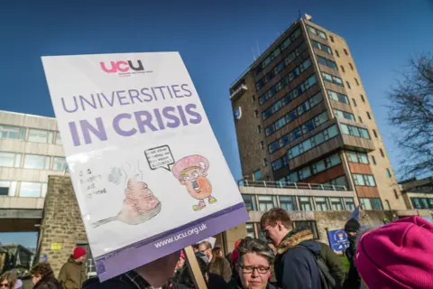 BBC A protester holds a placard reading “Universities in Crisis” with the UCU logo during a demonstration outside a university building. Other people are gathered around under a clear blue sky.