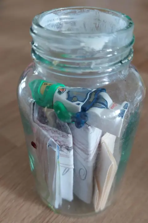Family The glass jam jar with its lid missing after it has been opened. The Lego spaceman is lying on the top of carefully-folded papers.