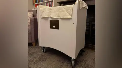 Chelsea Turrell A tall white cot which has wheels and has white blanket draped over it. It is standing in front of a fireplace and bookshelf. The room has a carpeted floor.