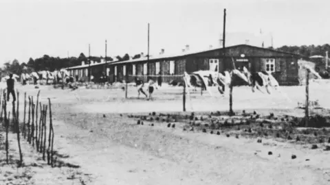 Keystone/Hulton Archive/Getty A grainy black-and-white photo of a prisoner-of-war camp, with men playing football in front of posts, washing hanging out and a series of single-story wooden huts.