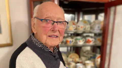 John is looking into the camera. He has very short grey hair and is wearing a black, cream and navy jumper with a black and white polka dot shirt on. He is standing in front of a wooden cabinet with glass doors that are open. Inside you can see dozens of brightly coloured ceramic coffee cups with matching saucers