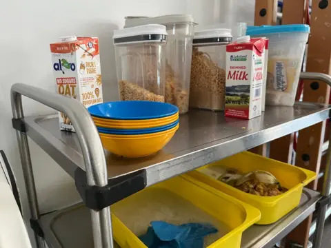 Blue and yellow plastic bowls are stacked the top shelf of a metal caddy, with cereal decanted into clear tubs and cartons of oat and cows milk nearby. On the middle shelf, there are two yellow tubs, one filled with water and a blue cloth and one with leftover food in it. 