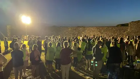 Walkers congregate at dawn for the annual Sunrise Walk of Hope across the Guernsey in aid of mental health. Some are wearing fluorescent jackets and the scene is lit by a bright spotlight. The castle walls are in the background.   