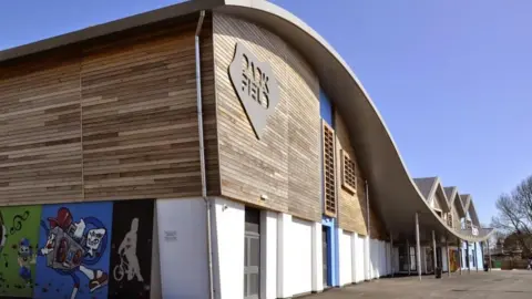 There are green, blue and black and white murals along the wall of a timber clad building with a sloping roof. The murals show a cyclist and a speaker playing music.