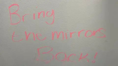 Anonymous An image of the words "Bring the mirrors back" written in pink highlighter on a bathroom wall