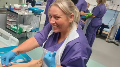 Rachel Ohene-Adjei Rachel wearing purple hospital scrubs and blue surgical gloves. She smiles at the camera and holds a syringe in one hand. 