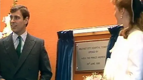 An older image shows a man with dark hair standing beside a commemorative plaque. He is wearing a grey pinstripe suit, a white shirt and green tie. A woman with red hair and wearing a white outfit is standing across from him with her back to the camera.