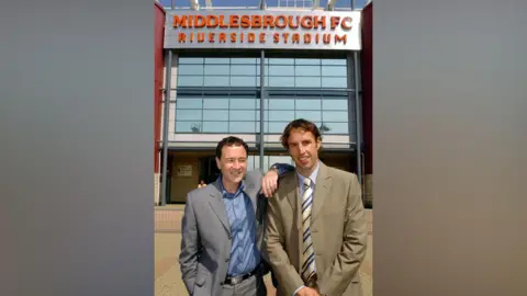 PA Media Gareth Southgate (right) and Steve Gibson stand in front of the red and silver façade of Middlesbrough FC's Riverside Stadium and are smiling at the camera. Gibson is wearing a grey suit and has his arm on Southgate's shoulder. Southgate is wearing a blue suit. The picture was taken in 2006.