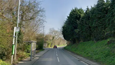A Google Streetview image of New Mill Lane, which is a tarmac road with one lane in either direction. There is a bus stop with metal and glass shelter to the left and a grassy verge and trees to the right