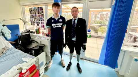 BBC Hamish (on the left wearing a dark blue sports top and shorts with his prosthetic legs) and Craig Mackinlay (on the right wearing a short, tie and blazer with his prosthetic legs) stand in a hospital room