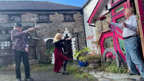 Sally Thomson Three people, two with model guns in their hands, pretending to shoot a man outside a country style pub.