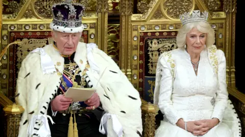 PA Media King Charles is sat down in a large gold chair next to Queen Camilla. Both of them are wearing crowns and fur outfits