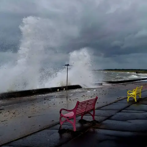 Angela Martin Tall waves crash against the barriers at Prestwick Beach. It appears to be a windy day with dark storm clouds. Two benches, one red and one yellow, sit back from the water.