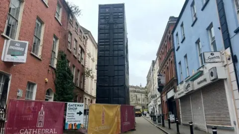 large shipping containers stand upright in a narrow pedestrianised street. Shops line both sides of the street. A number of crash barriers are in front of the container.