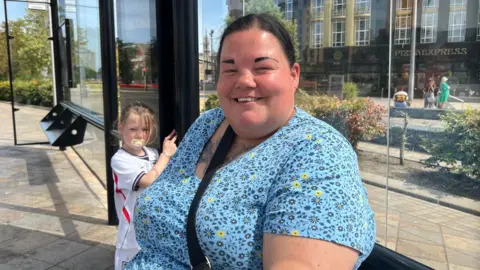 A woman wearing a blue dress is sitting in a bus shelter with her young daughter who is wearing an England football shirt. A public square and a smart-looking restaurant can be seen in the background.