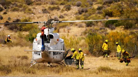 Firefighters in yellow hi vis and helmets gather around a helicopter parked on shrubland in Almocaizar on 12 July.
