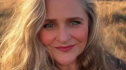 Headshot of a blonde curly haired woman with blue eyes and a nose ring, standing in a field