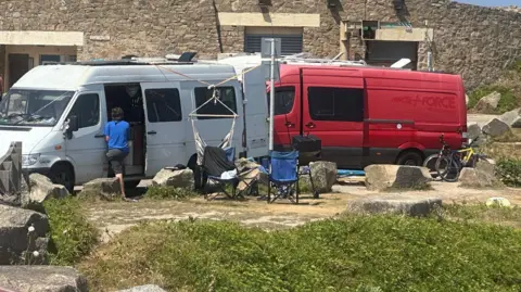 A coastal car park and road, with numerous vans, campervans and cars parked up.