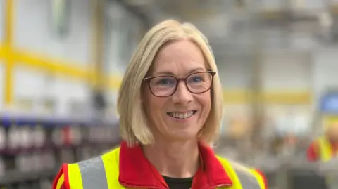 Julie Thomas is a woman with blonde, short hair and glasses. She is wearing a red fleece with a high-viz vest. She is smiling at the camera. The background is an out-of-focus postal sorting office.