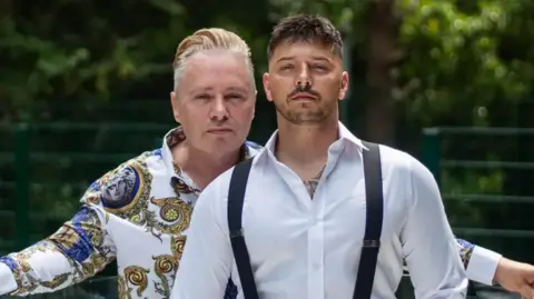 Barrie Drewitt-Barlow On the left is Barrie, looking towards the camera. He has blonde hair and a gold and blue shift on. On the right is Scott, who is wearing a white shirt and black braces. He has brown hair and is also looking towards the camera. 