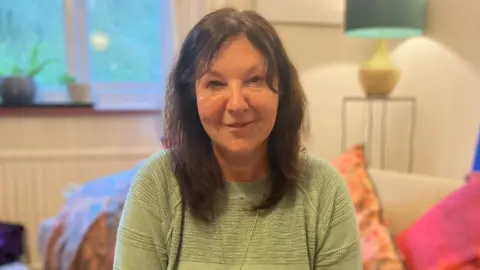 BBC/Simon Dixon-French A dark-haired woman in a light green top. She is sitting on a sofa with several colourful pillows. 
