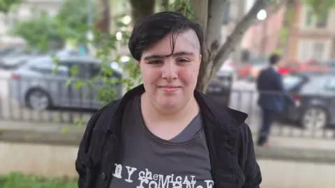 BBC Girl with black hair standing by a tree, smiling.