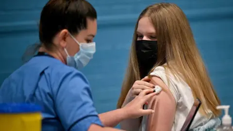 A health worker wearing a blue shirt and blue face mask holds a small cotton pad on another woman's shoulder. The other woman has long blonde hair, a black face mask and a white top.